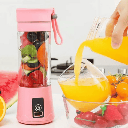 Pink portable blender with fruits inside, next to a glass of orange juice and more fruits on a white background.