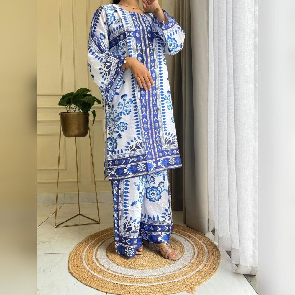 Person wearing a blue and white patterned outfit standing in a room with a plant and curtain.