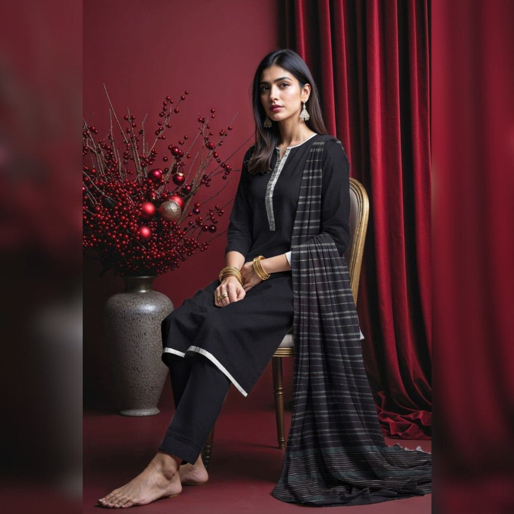 Woman in black traditional outfit sitting on a chair with a red curtain and decorative plant in the background