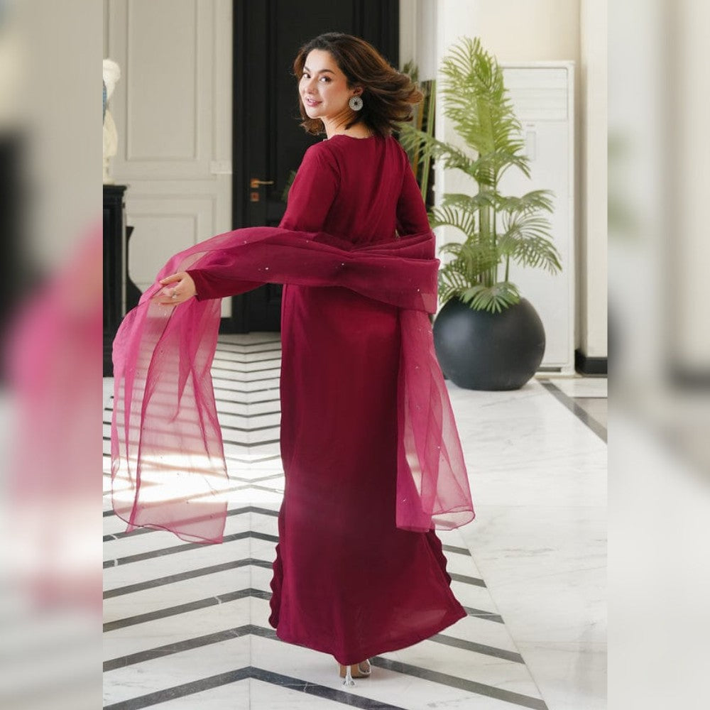 Woman in a burgundy dress with a sheer overlay standing in an elegant interior setting.