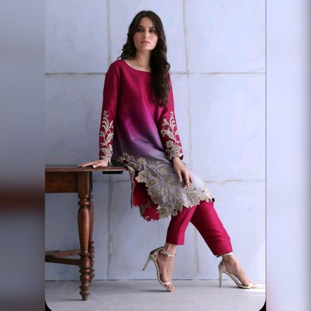 Woman in a magenta outfit with gold embroidery sitting on a wooden stool against a light-colored wall.